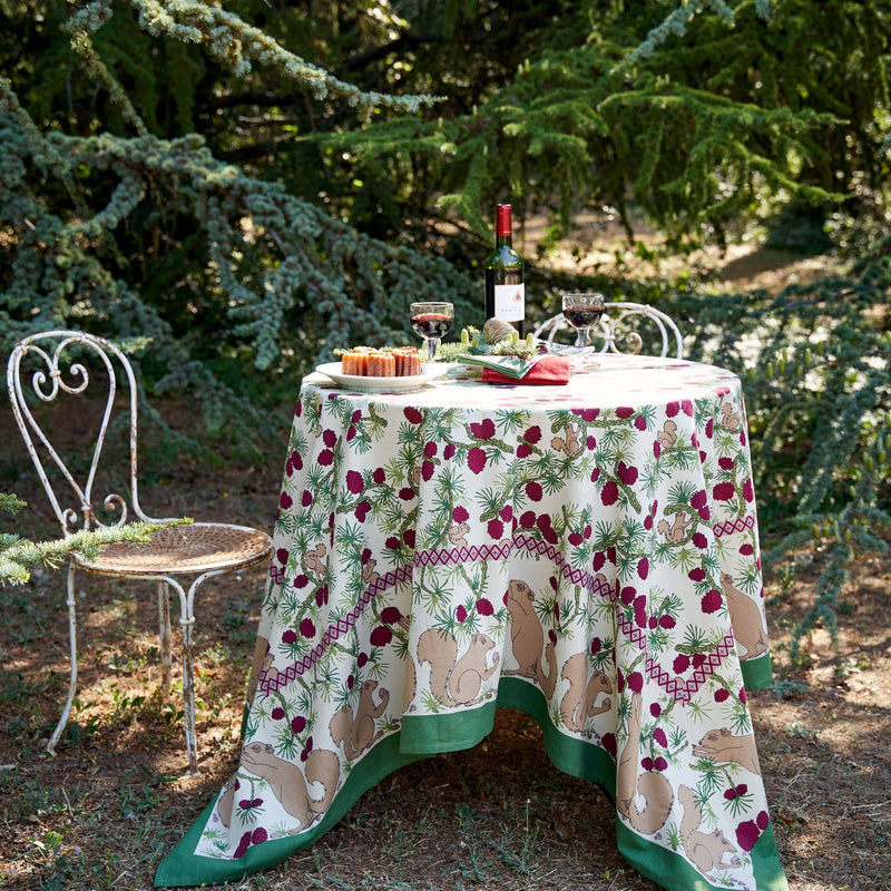 Couleur Nature - Squirrel and Pinecone Red & Brown Tablecloth: 71 x 71 drapes a woodland bistro table with wine, pastries, and evergreen decor in a rustic garden setting.