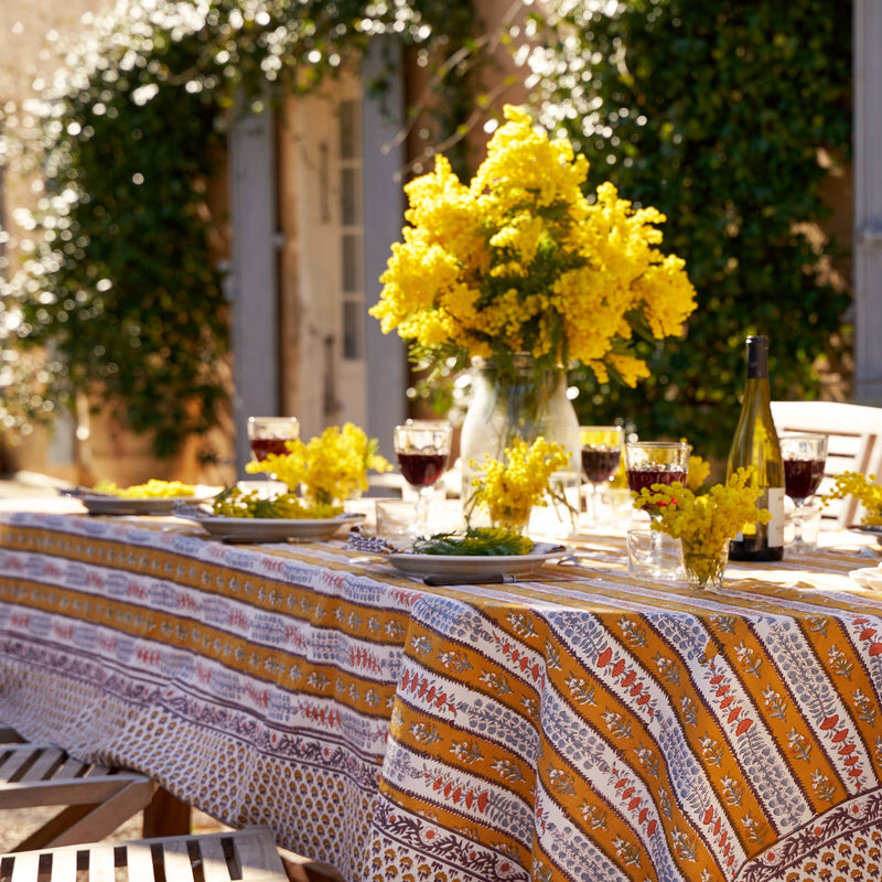 Couleur Nature - Provence Avignon Dijon & Blue Tablecloth: 71 x 71 on an outdoor dining table, adorned with mimosa flowers, place settings, and wine glasses in a sunlit garden.