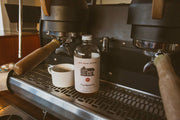 Pink House Alchemy - Cardamom Simple Syrup bottle next to a coffee machine, highlighting its perfect pairing with coffee, ideal for enhancing beverages.