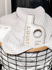 L'AVANT Collective - Laundry Detergent Fresh Linen bottle atop a black wire basket of white waffle towels, embodying eco-friendly laundry care.