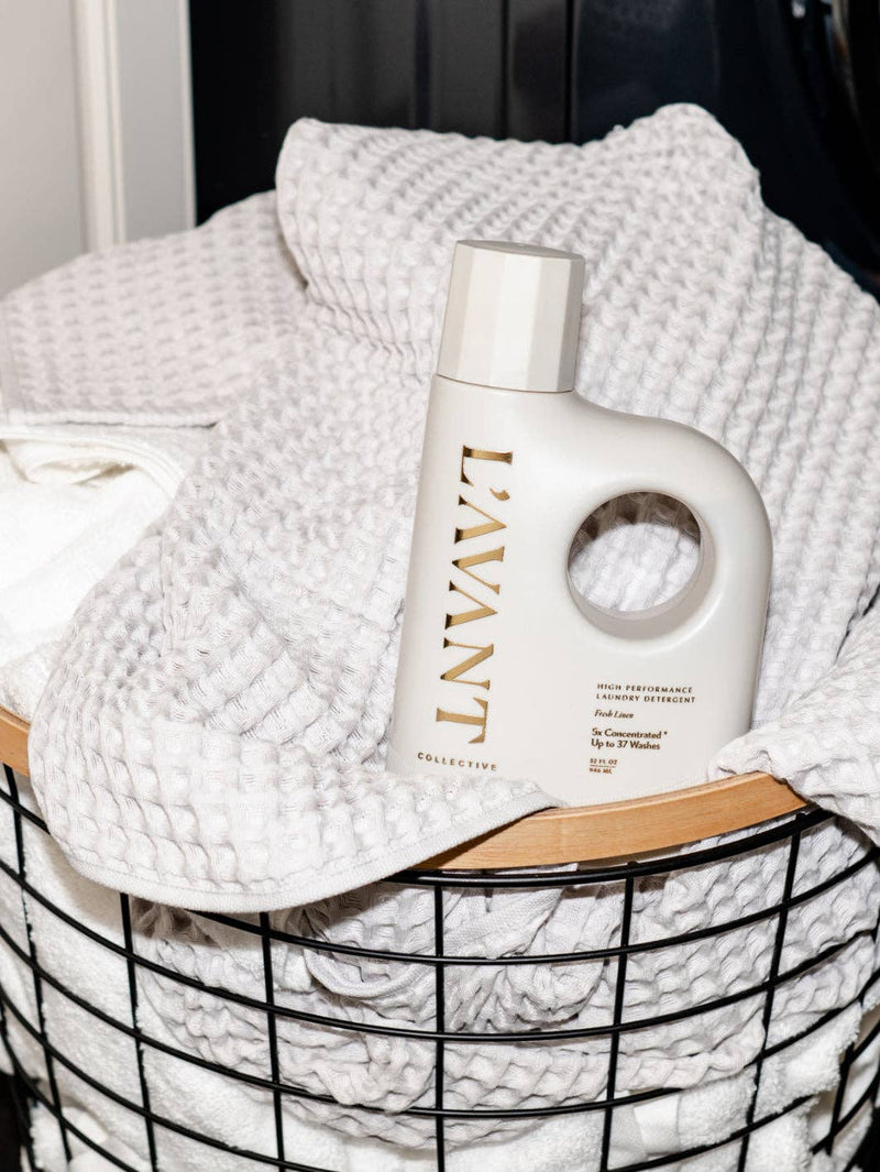 L'AVANT Collective - Laundry Detergent Fresh Linen bottle atop a black wire basket of white waffle towels, embodying eco-friendly laundry care.