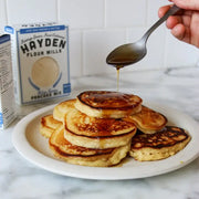 Hand drizzles syrup onto fluffy pancakes made with Hayden Flour Mills - White Sonora Pancake Mix, showcasing its minimal-ingredient, buttery grain goodness on a marble countertop.