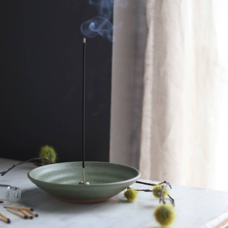 Gravesco Pottery Incense Burner holds a glowing incense stick, surrounded by matches, a glass vial, and spiky seed pods, creating a serene, contemplative setting.