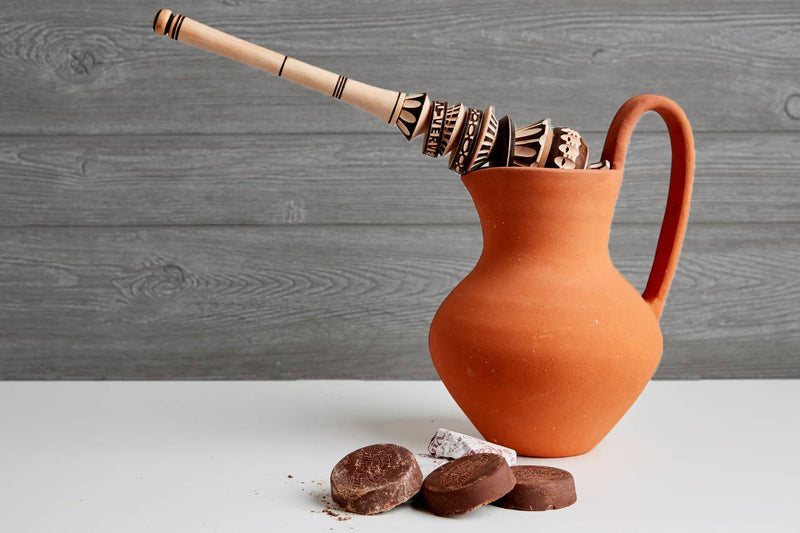 Villa Real Mexican Hot Chocolate: Almond / Large - 10 Discs displayed with terracotta pitcher and wooden tool on a white surface with cocoa dust.