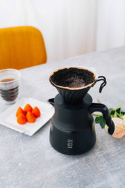 HARIO USA - V60 Metal Dripper: Silver atop black carafe, with coffee cup and strawberries nearby, showcasing precision brewing setup.
