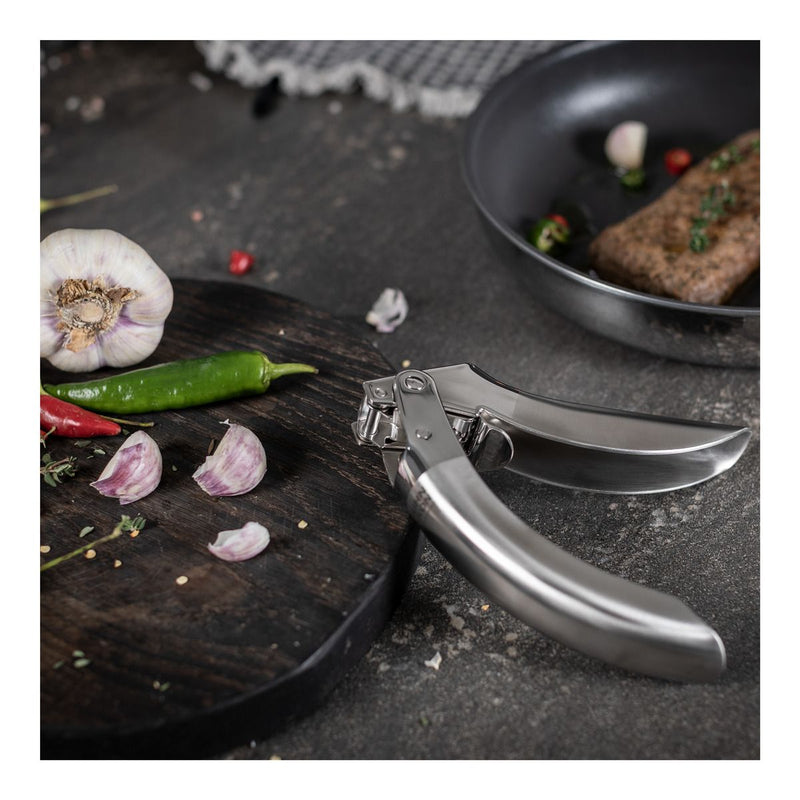 Kuhn Rikon | Epicurean Garlic Press on countertop beside cutting board with garlic, chilies, and herbs; ergonomic stainless steel design with easy-clean pullout sieve.