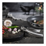 Kuhn Rikon | Epicurean Garlic Press on a cutting board with garlic and chili peppers, showcasing its stainless-steel design and ergonomic handle for effortless garlic pressing.