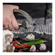 Kuhn Rikon | Epicurean Garlic Press minces garlic effortlessly onto a plate, surrounded by whole cloves and herbs, showcasing its ergonomic, stainless-steel design and easy-to-clean features.