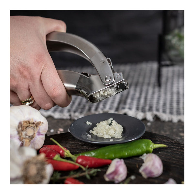 Kuhn Rikon | Epicurean Garlic Press minces garlic effortlessly onto a plate, surrounded by whole cloves and herbs, showcasing its ergonomic, stainless-steel design and easy-to-clean features.