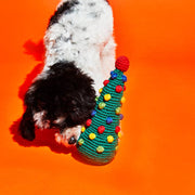 Ware of the Dog - Pom Pom Tree toy, a hand-knit, cone-shaped plaything with colorful pom-poms, being investigated by a fluffy puppy.