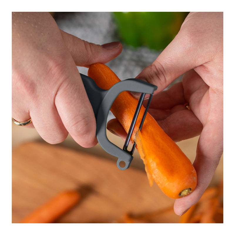 Kuhn Rikon | Original Swiss Peeler - Grey held over a cutting board, peeling a carrot with its sharp carbon steel blade in a close-up view.