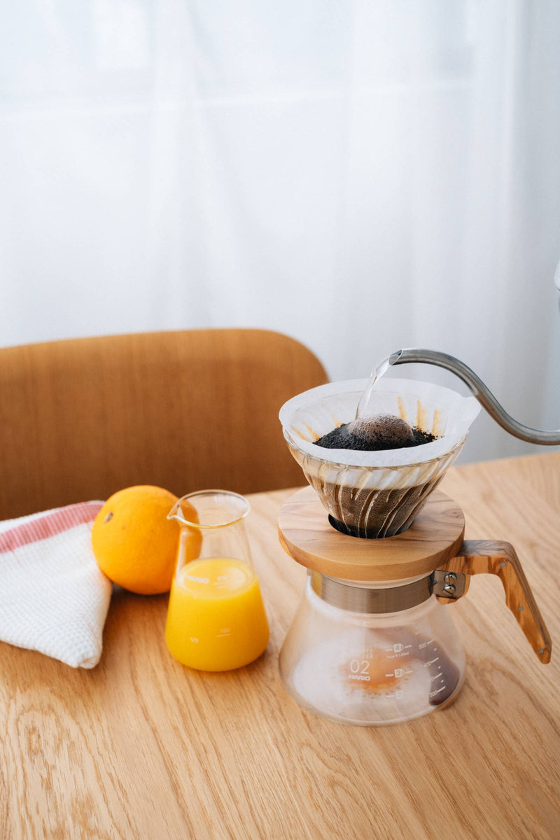 HARIO V60 Glass Dripper 02 with olive wood holder pouring coffee, alongside orange juice and fruit on a kitchen table.