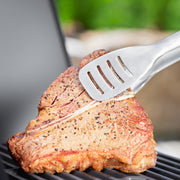Rösle | Premium Barbecue Grilling Tongs precisely turning steak on a grill, showcasing large turning surface and grease drain slots for efficient cooking.