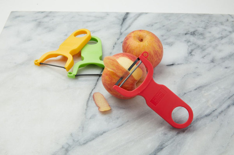 Kuhn Rikon | Original Swiss Peeler Set with three Y-shaped peelers on marble, featuring a red peeler actively peeling an apple.