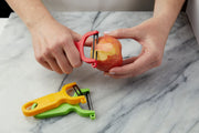 Hands using Kuhn Rikon Original Swiss Peeler in red on an apple; yellow and green peelers beside, demonstrating ergonomic design and sharp blades.