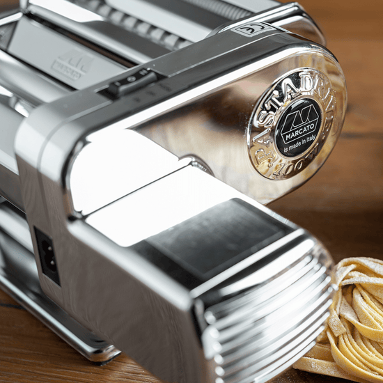 Marcato Atlas PastaDrive Motor 110V Premium on wooden surface, showcasing chrome-plated housing and dough thickness adjustment knob, with freshly cut, flour-dusted pasta beside it.