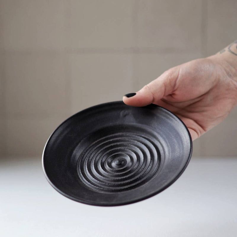 Gravesco Pottery - Garlic Grater Plate in Black, hand holding a matte-black ceramic dish with concentric ridges, ideal for garlic preparation and serving.