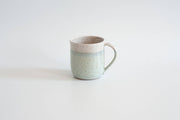 RachaelPots - 12 Oz Original Mug: Sage, a handmade ceramic mug with a mint-green speckled glaze and beige band, featuring a thumb rest handle. Perfect for coffee or tea.