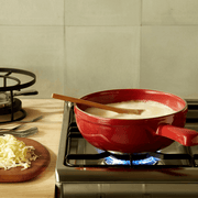 Emile Henry | Cheese Fondue Set, Burgundy, shown melting cheese over a gas burner with fondue forks and shredded cheese on a wooden board nearby.