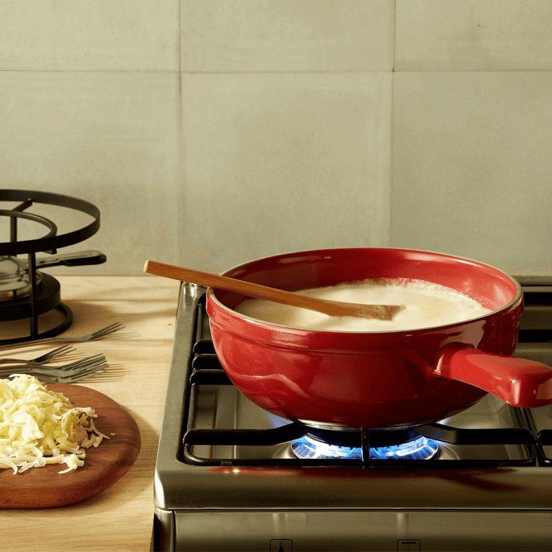 Emile Henry | Cheese Fondue Set, Burgundy, shown melting cheese over a gas burner with fondue forks and shredded cheese on a wooden board nearby.