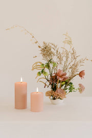 Mo&Co Home - 100% Beeswax Pillar Candles | Rose Pink: 3x4 Inch. The image shows two pink beeswax candles alongside a vase with flowers, highlighting their elegant design.