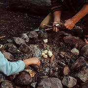 Bee's Wrap - ReKindle Fire Starters™ being used in a stone-ringed campfire with hands arranging sticks and roasting items on embers.