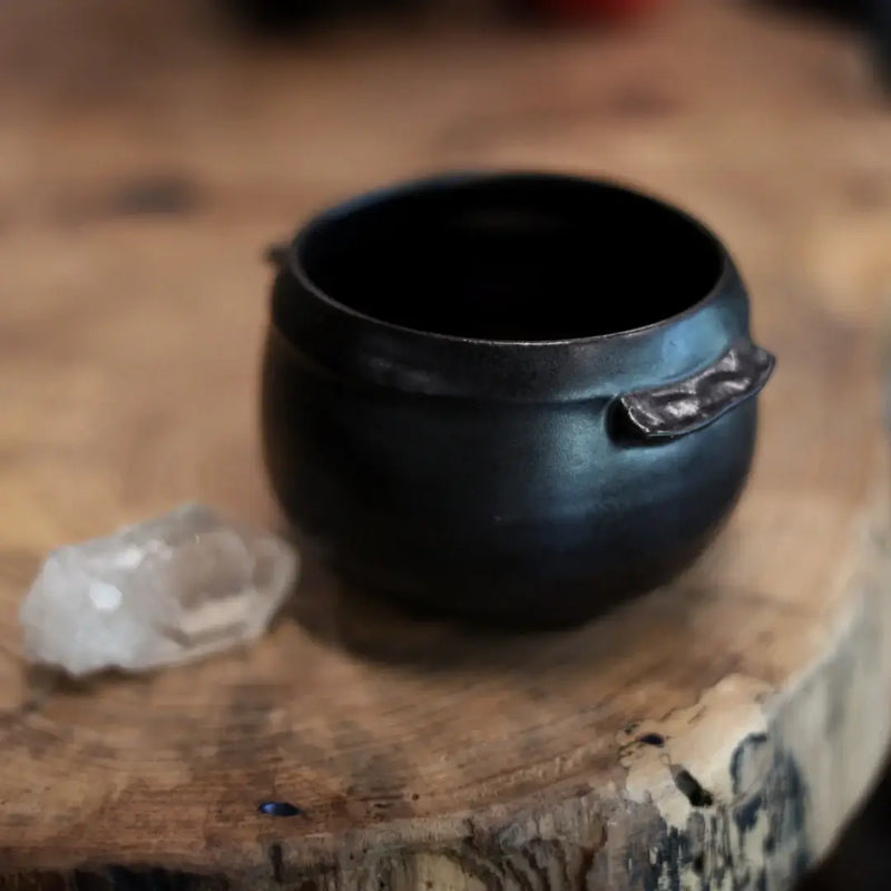 Gravesco Pottery - Black Cauldron Bowl, handcrafted with rustic charm, sits on wood beside a quartz crystal, embodying artisanal craftsmanship and magical, apothecary style.