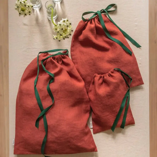 MagicLinen | Christmas Gift Bags, Set of 3: Three burnt-orange linen drawstring bags with dark green ribbons, perfect for reusable gift-giving or organizing items.