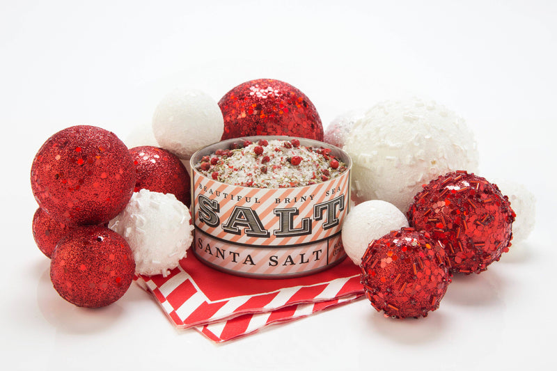 Beautiful Briny Sea - Santa Salt tin on striped napkins, surrounded by festive decorations, highlighting its use in enhancing various dishes with its unique blend.