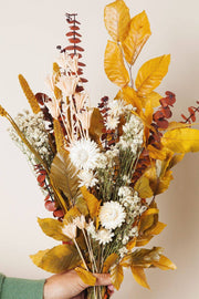 Idlewild Floral Co. - Golden Fall Market Bouquet: A hand holds a warm, autumnal dried-flower arrangement with yellow leaves, red eucalyptus, and white daisies.