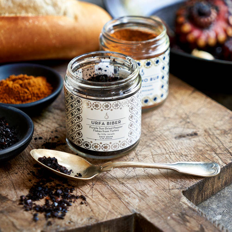 Villa Jerada - Urfa Biber - 50g jar on wooden board, with spoon displaying pepper flakes, surrounded by spice jars and bread, embodying complex, smoky, mildly spicy flavor.