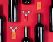 L'Atelier du Vin - Twin Blade Wine Opener, BILAME CRAFT, with tan leather sheath, showcased among wine bottles, highlighting its artisanal craftsmanship and unique cork extraction method.