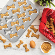Nordic Ware | 2 Piece Puppy Love Treat Pan and Dog Treat Mix alongside a tray of bone-shaped dog treats and a bowl of peanut butter.