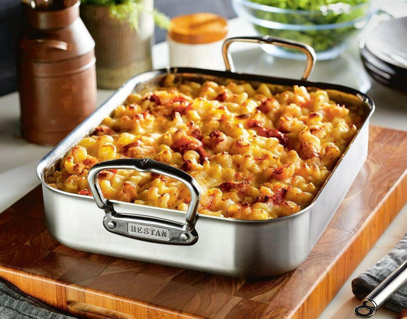 Hestan Provisions Classic Roaster with Rack, filled with baked macaroni-and-cheese, displayed on a cutting board. Ideal for versatile cooking and serving.