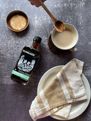 Runamok - Peppermint Bark Organic Maple Syrup 250ml beside a frothy coffee, with a hand poised to sprinkle cinnamon, highlighting a cozy holiday treat.