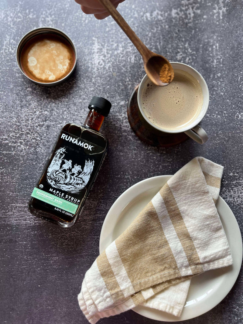 Runamok - Peppermint Bark Organic Maple Syrup 250ml beside a frothy coffee, with a hand poised to sprinkle cinnamon, highlighting a cozy holiday treat.