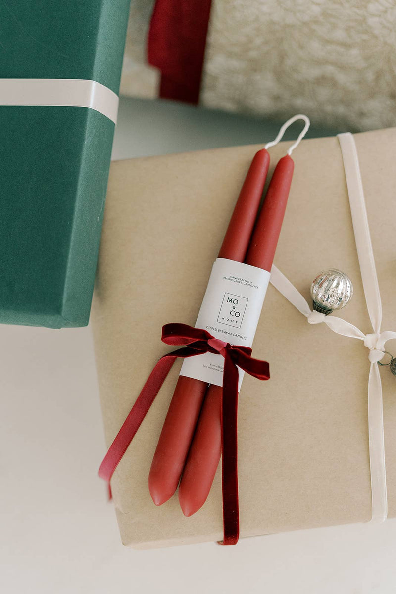 Mo&Co Home - Berry Red | Dipped Beeswax Taper Candles, paired and velvet-tied, rest atop elegantly wrapped gifts, embodying natural elegance and sustainable craftsmanship.