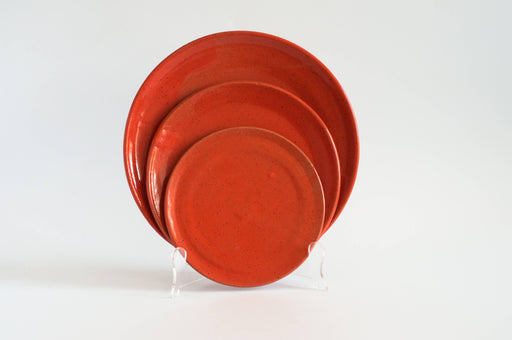 RachaelPots - Dinner Plate: Coral, set of three red ceramic plates, nested on a clear stand. Hand-thrown stoneware with an upturned lip, crafted in Brooklyn.