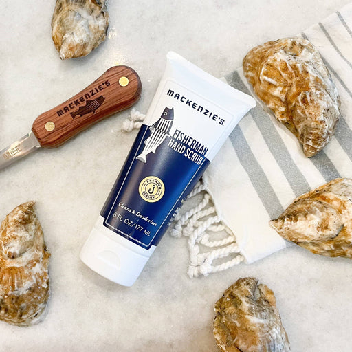 Mackenzie's Fisherman Hand Scrub 6 fl oz tube, surrounded by oyster shells, a branded oyster knife, and a kitchen towel, on a marble countertop.