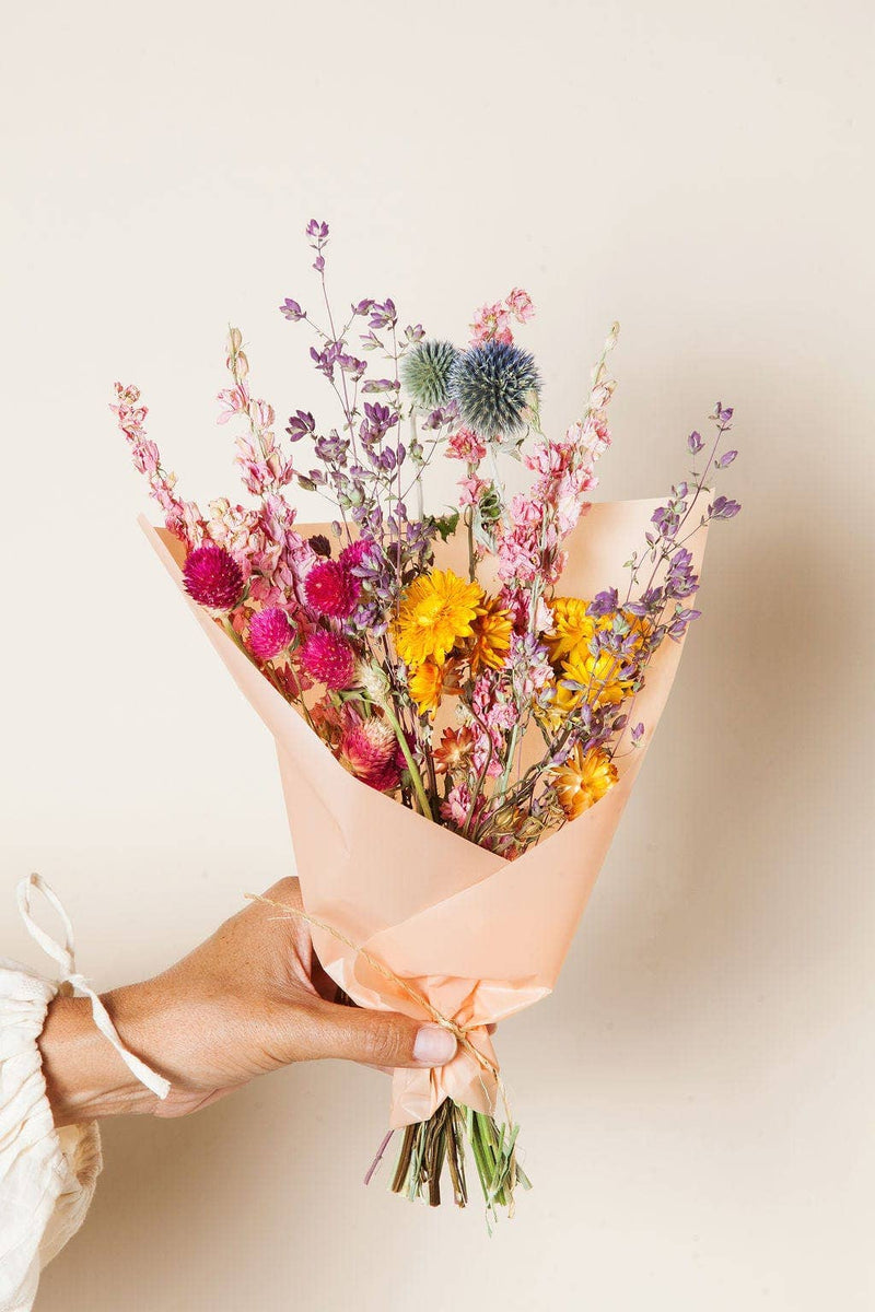 Idlewild Floral Co. - Bright Small Field Bouquet: Hand holding a dried wildflower bouquet with strawflowers, globe amaranths, and thistle-like blooms, wrapped in peach paper.
