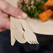 Hand holding a bambu® Disposable Bamboo Knife, Fork, and Spoon Set on an eco-friendly bamboo plate with salad and roasted vegetables, showcasing sustainable dining.