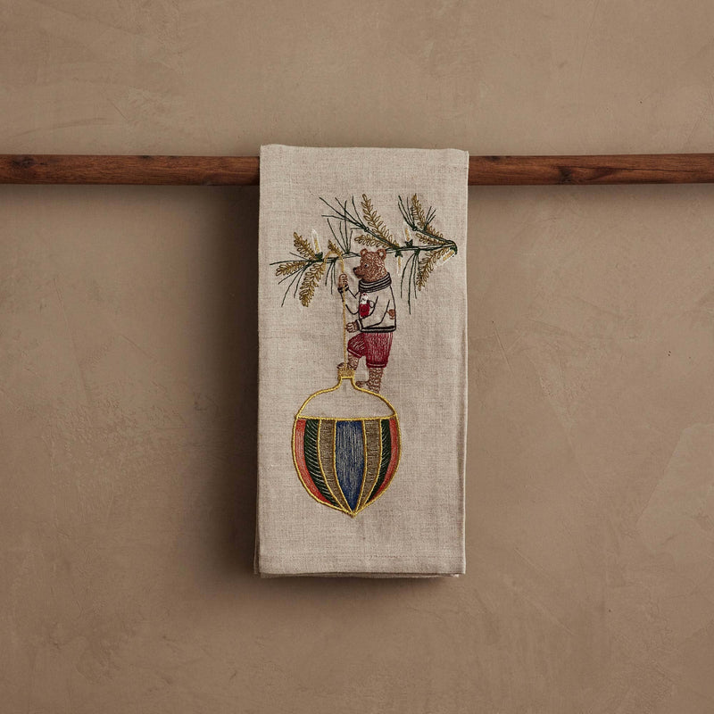 Coral & Tusk - Deck the Halls Bear Tea Towel featuring an embroidered bear on evergreen branches with a striped ornament, draped over a wooden rod.