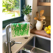 Bespoke Provisions - Tropical Leaves Swedish Dishcloth displayed on a kitchen sink handle, accompanied by lemons, a plant, and a pitcher with utensils, highlighting its kitchen utility.