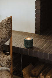 Emerald Green Marble Refillable Candle in textured holder on a brick hearth, alongside sheepskin-draped chair, exuding cozy warmth with stacked firewood below.