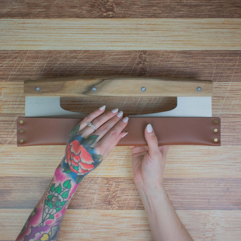Tattooed hand holding Lamson leather sheath for 14 pizza cutter/rocker, highlighting craftsmanship and durability, ideal for kitchen enthusiasts.