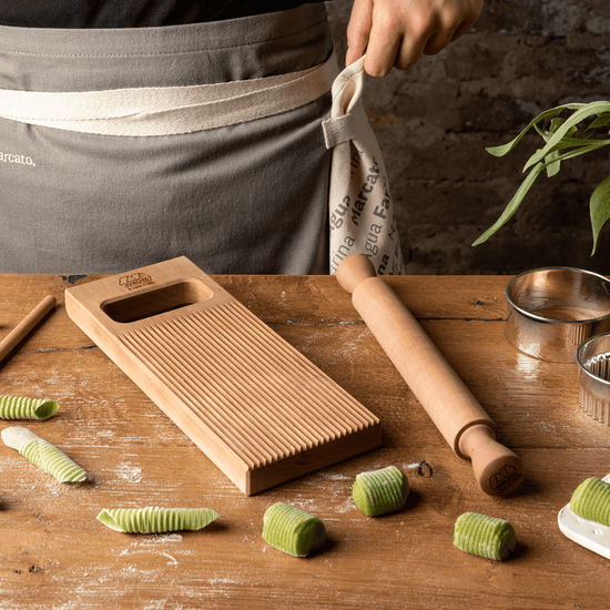 Marcato | Gnocchi Like A Pro Kit: Gnocchi board, rolling pin, and gnocchi-shaped dough on a tabletop, with apron and pastry rings in view.