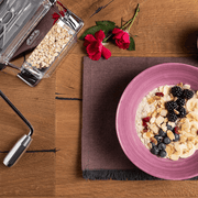 Top-down view of a breakfast scene featuring the Marcato | Mill + Grain Crusher with rolled oats beside a vibrant oatmeal bowl topped with assorted fruits and nuts.