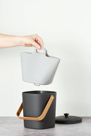 Hand holding the Bamboozle - Composter Reusable Liner above a black bucket, showcasing its grip loop for easy waste disposal.