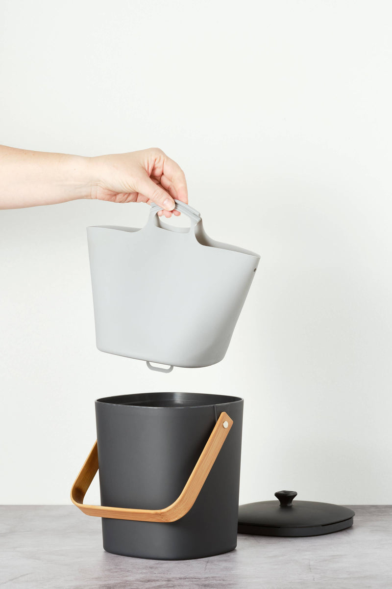 Hand holding the Bamboozle - Composter Reusable Liner above a black bucket, showcasing its grip loop for easy waste disposal.
