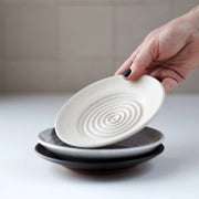 Gravesco Pottery - Garlic Grater Plate in Black. A hand lifts an off-white grater plate with concentric circles, ideal for preparing garlic and serving dips.